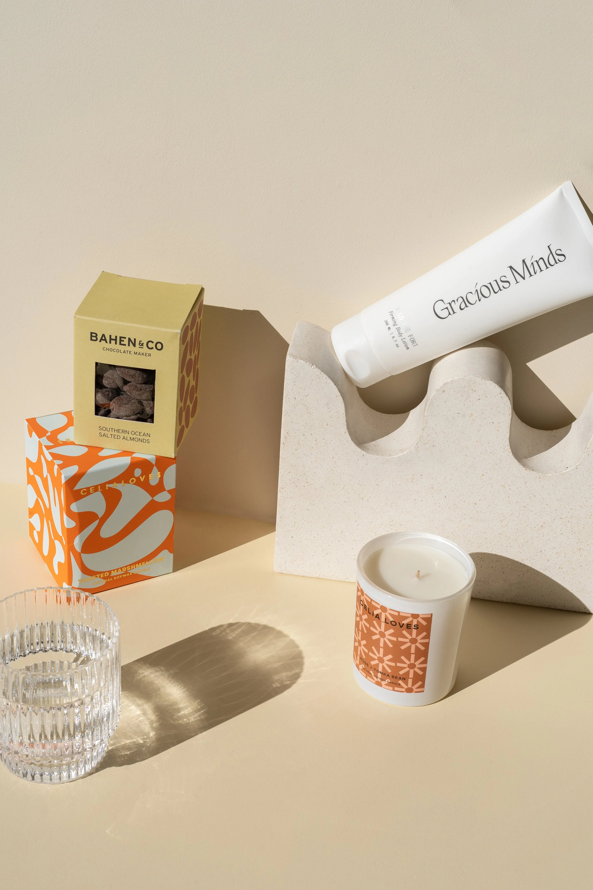 A minimalist still life of skincare products, candles, and snacks on a beige surface with shadows.