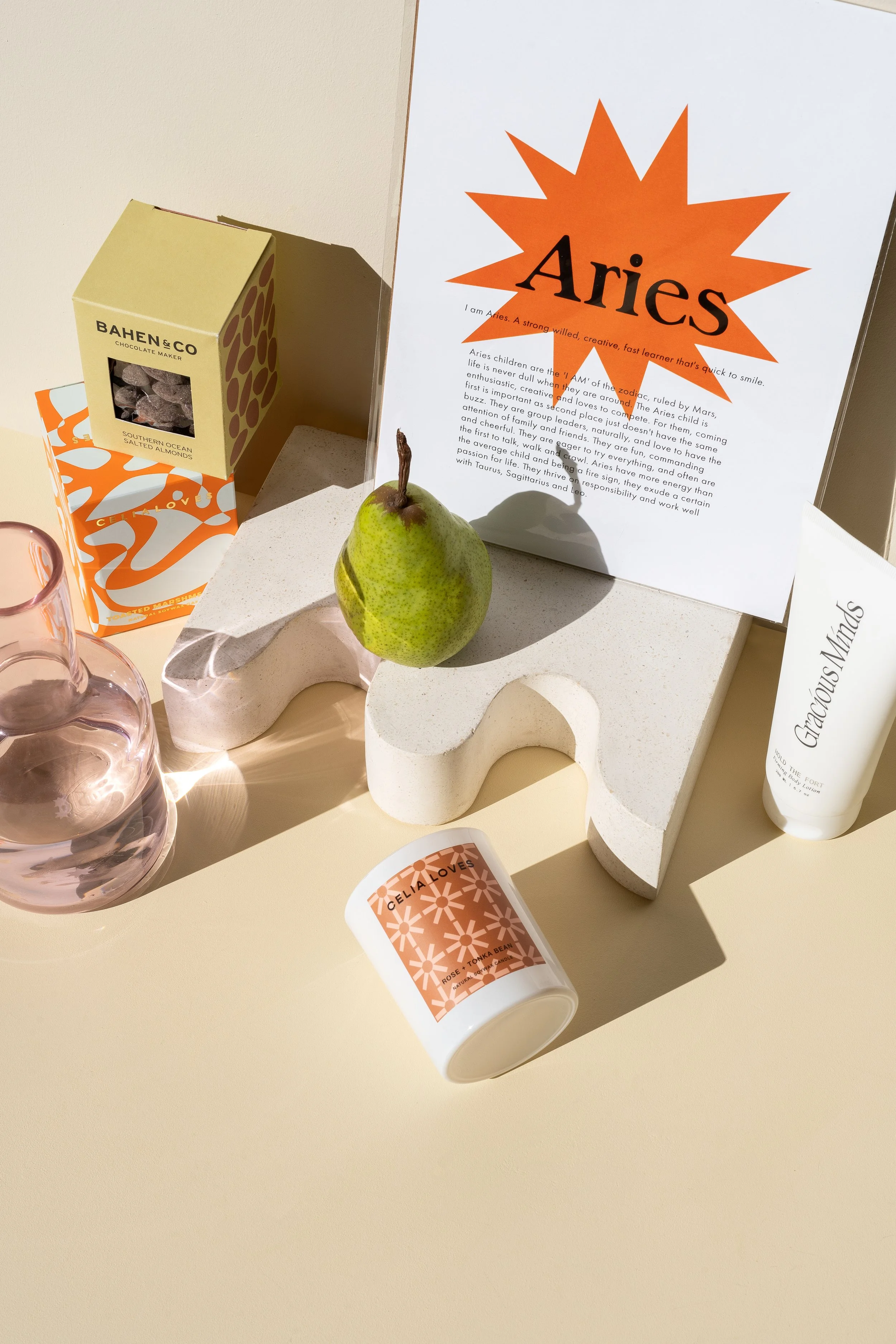 A collection of assorted items on a light yellow surface, including a pink glass bottle, a green pear, a candle labeled 'CELIA LOVES,' a container of 'BAHEN & CO' salted almonds, a bottle of 'Gracious Minds' lotion, and a framed paper with the word 'Aries' in bold orange lettering.