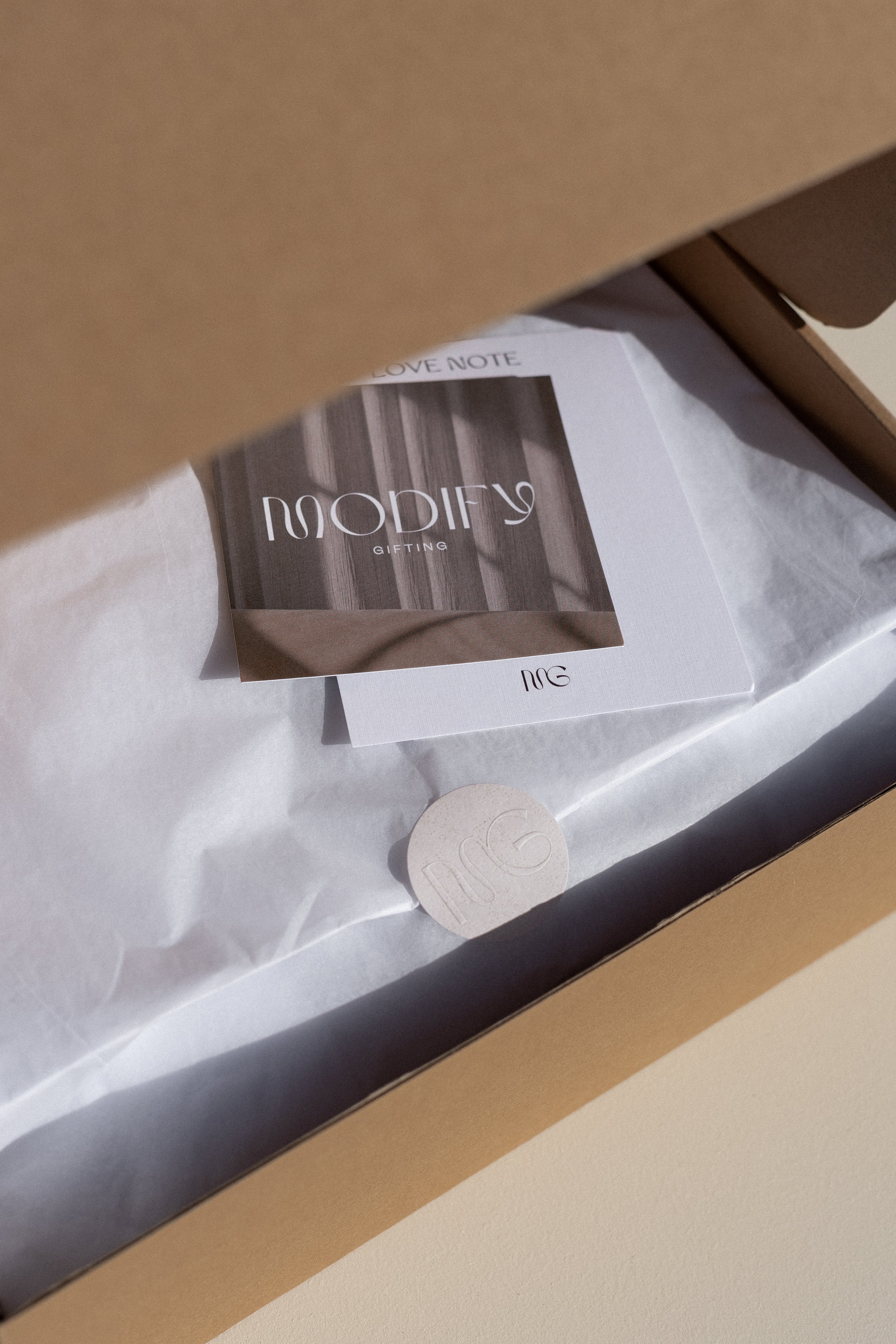 Open cardboard box with tissue paper, a card and a round item inside.