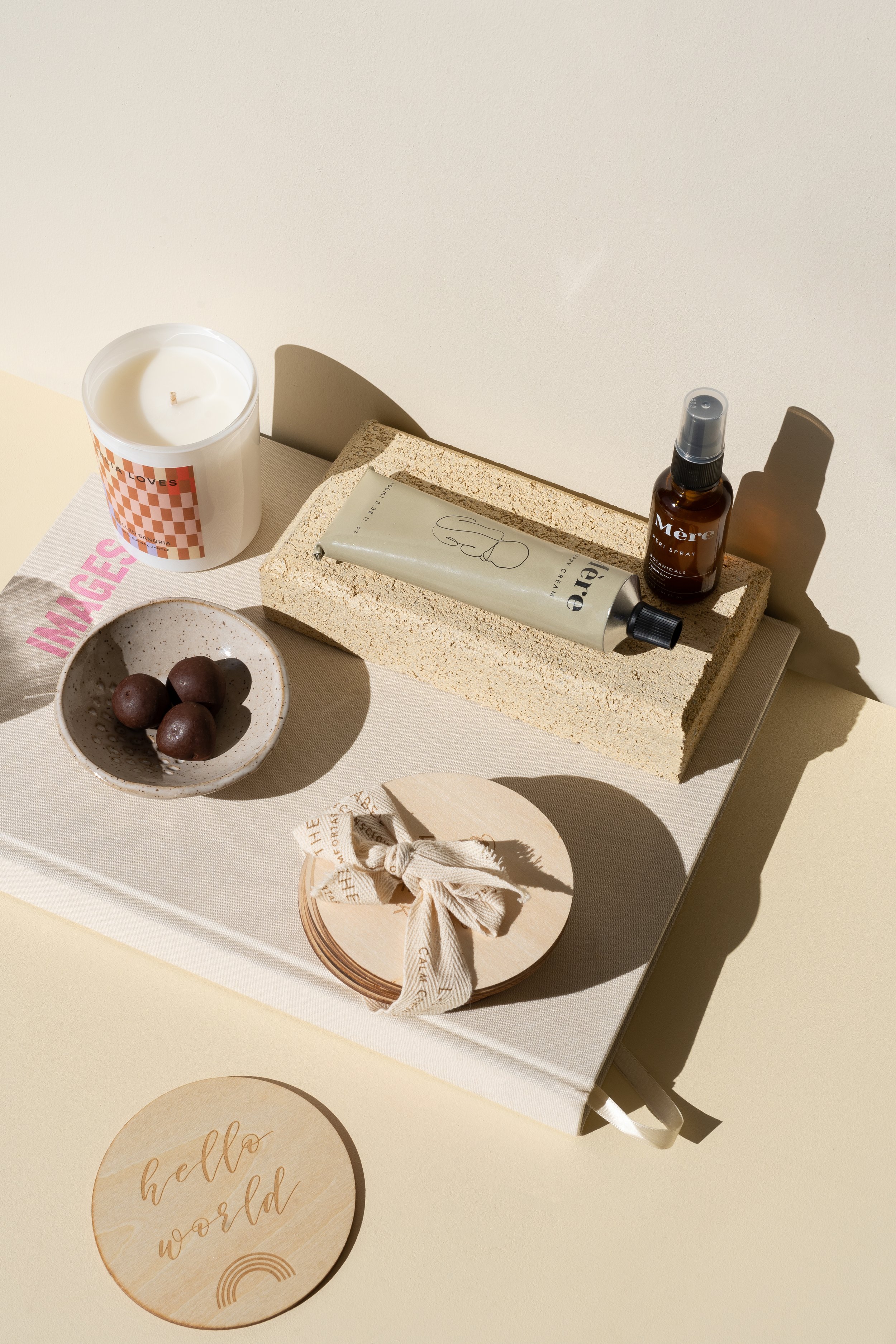 A beige tray with skincare products, a small bowl of chocolate balls, a candle, a wood coaster with a fabric bow, and a wooden coaster with the words "hello world" written on it.