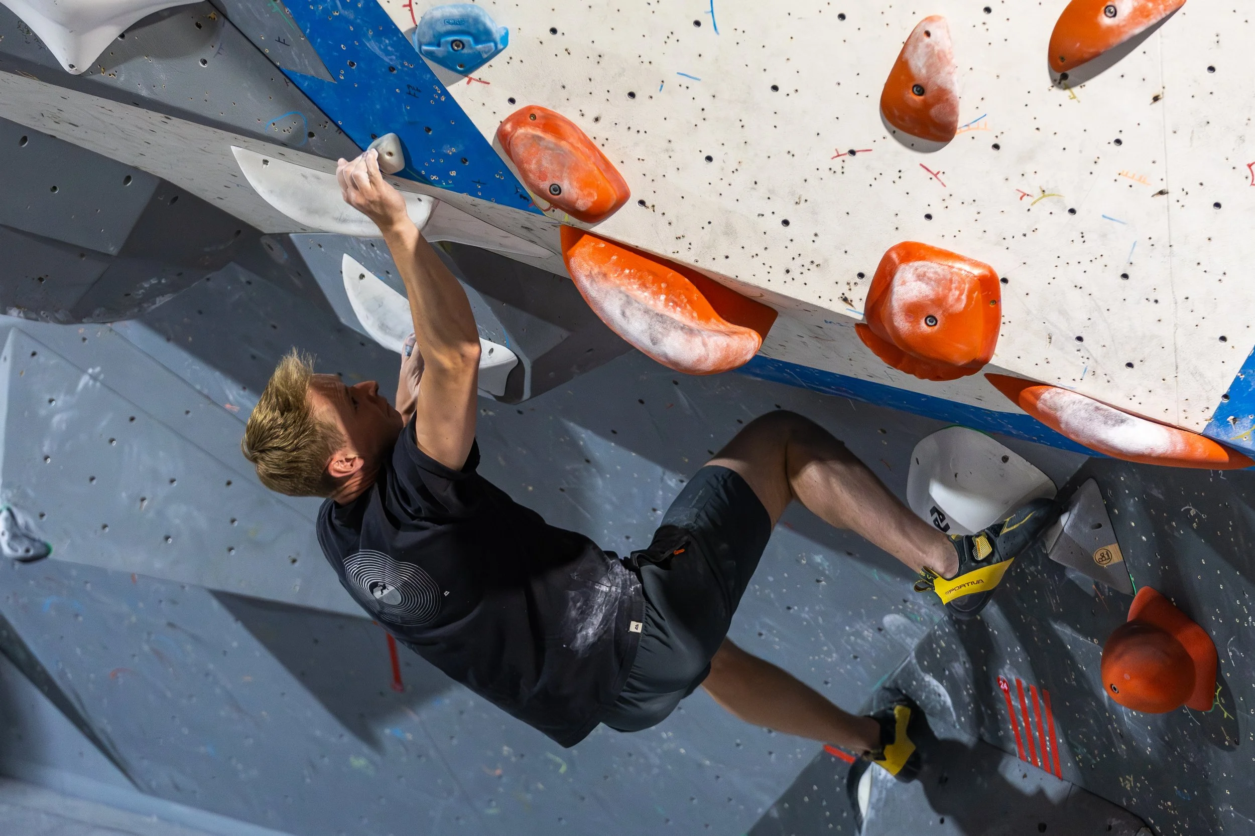 A person with blond hair rock climbing on an indoor climbing wall with orange holds, wearing a black shirt and yellow climbing shoes.