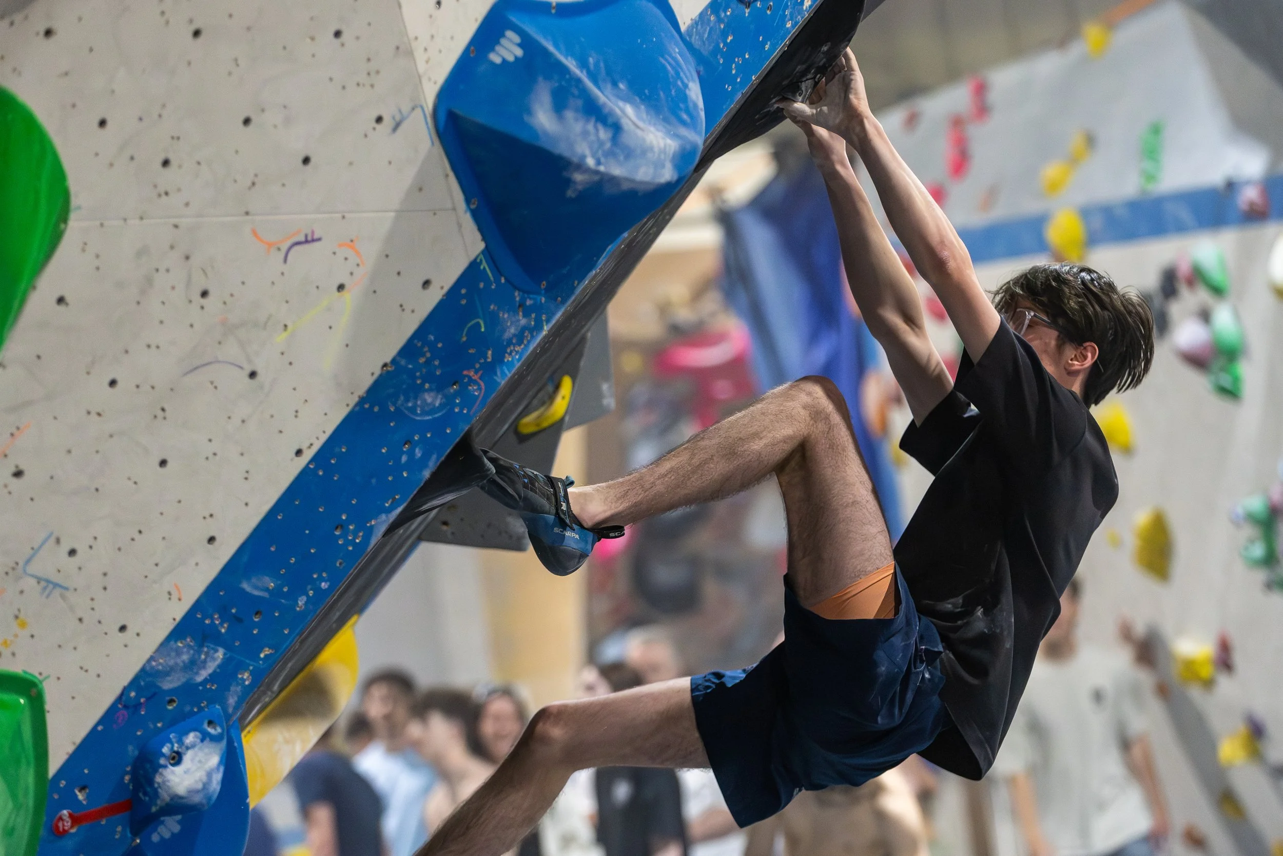 A person climbing an indoor rock wall, reaching for holds with an audience watching in the background.