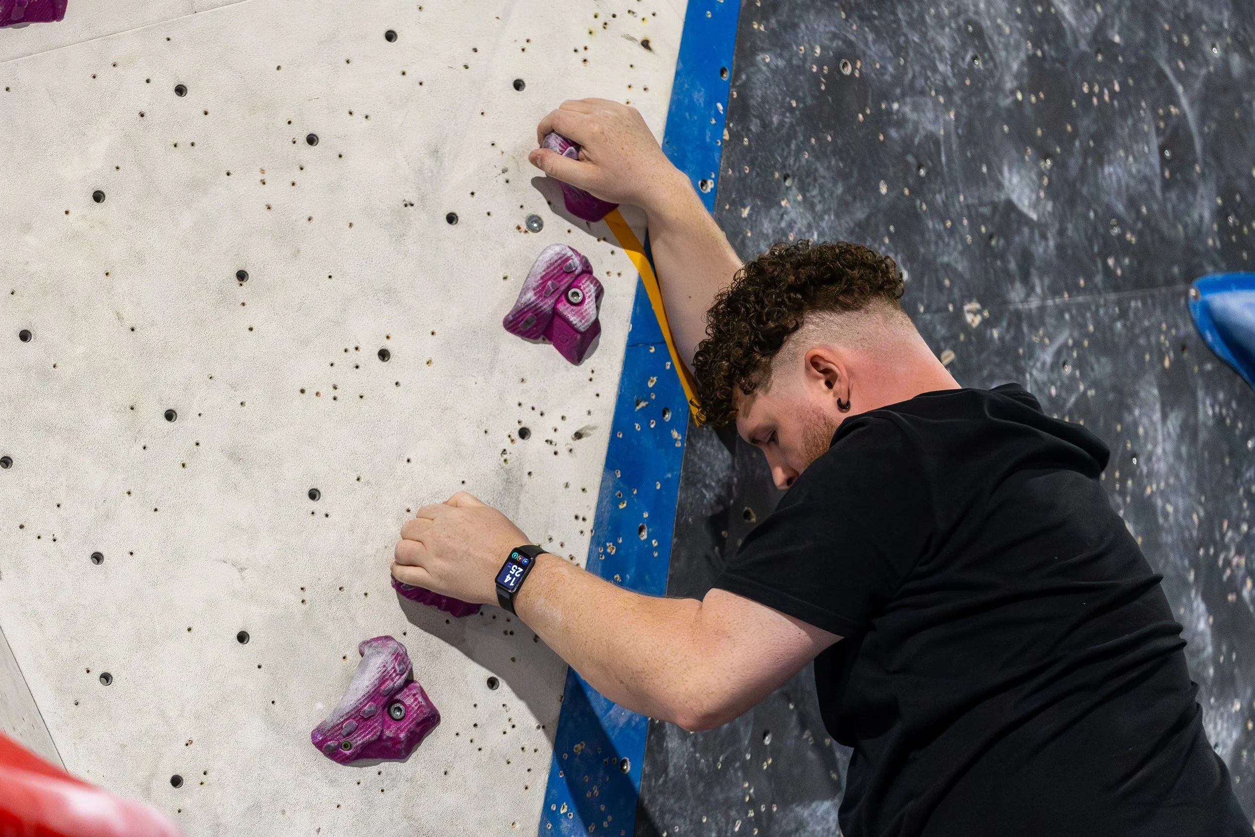 A person with curly hair and a black shirt is climbing an indoor rock wall. They are wearing a smartwatch on their left wrist and are gripping pink climbing holds on the wall, which is white with black bolt holes.