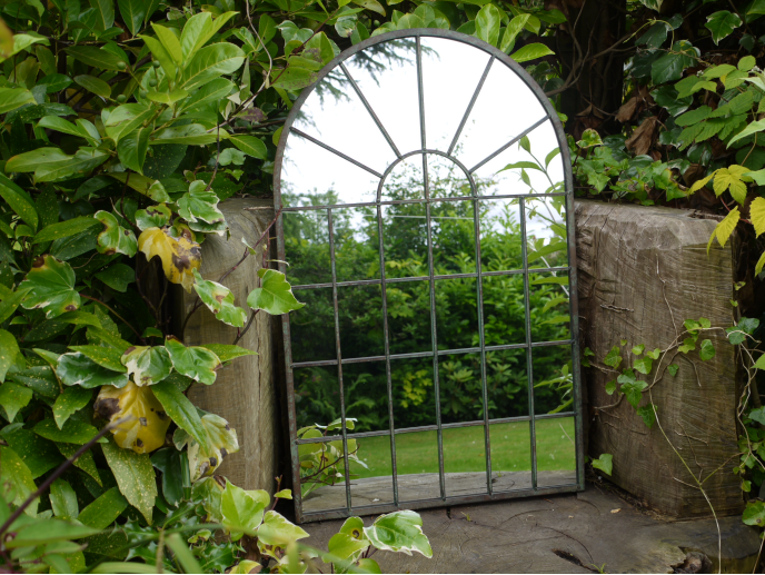 A decorative metal garden mirror with an arched top and grid design, leaning against a wooden fence, surrounded by green foliage.