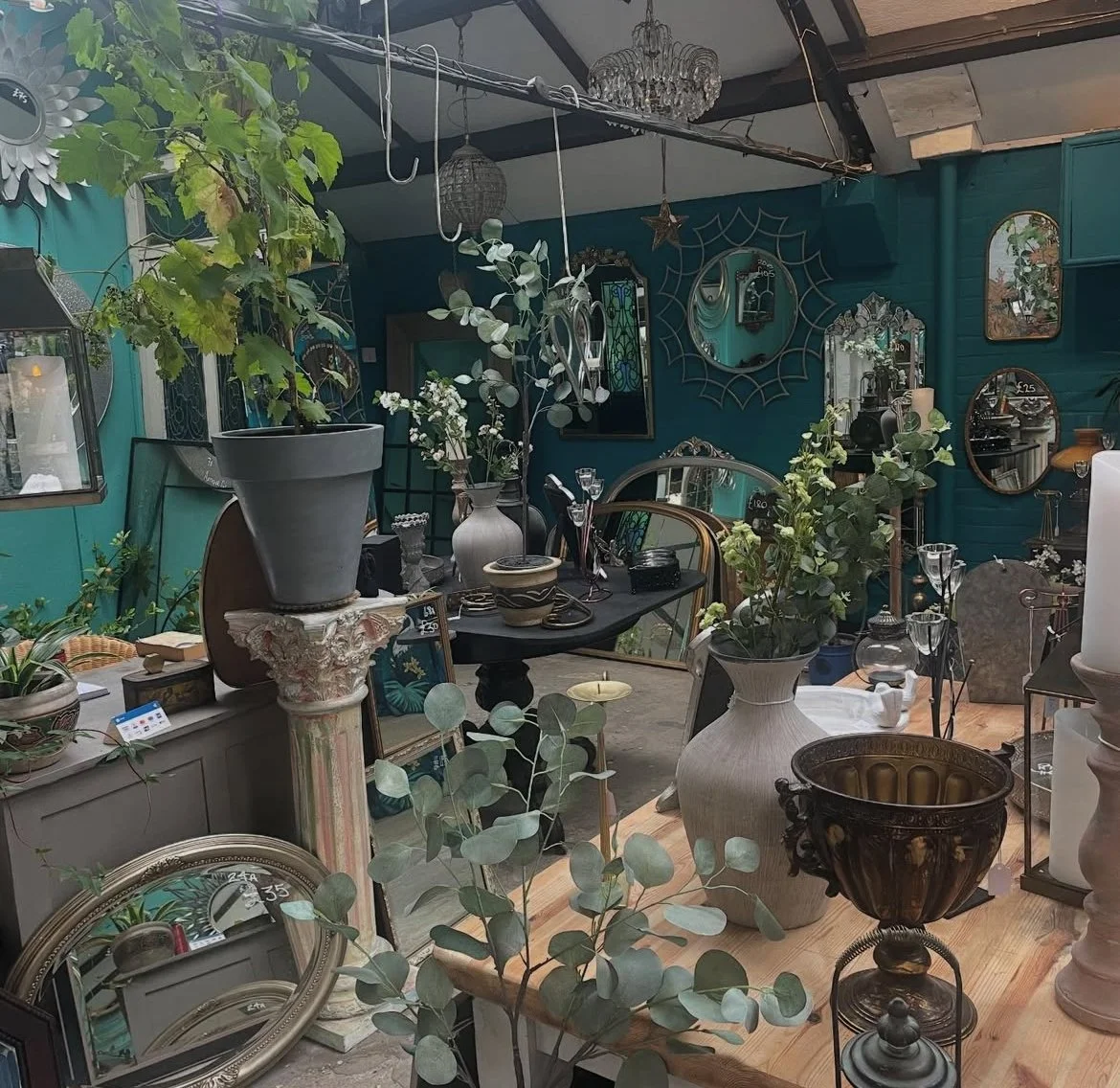Interior of a vintage store with various decorative items including vases, mirrors, framed pictures, plants, and chandeliers on a wooden floor and teal walls.