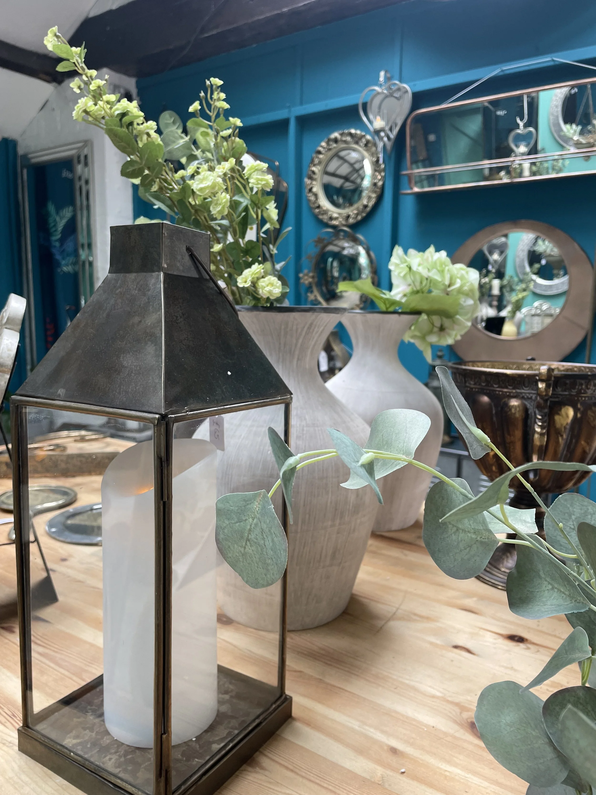 Decorative antiques and vases on a wooden table, including a lantern with a candle, surrounded by green plants, with a blue wall and mirrors in the background.