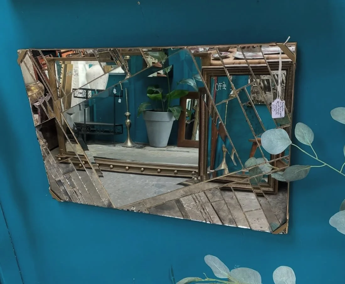 Decorative mirror with a gold geometric frame hanging on a teal wall, reflecting a potted plant, picture frames, and furniture.