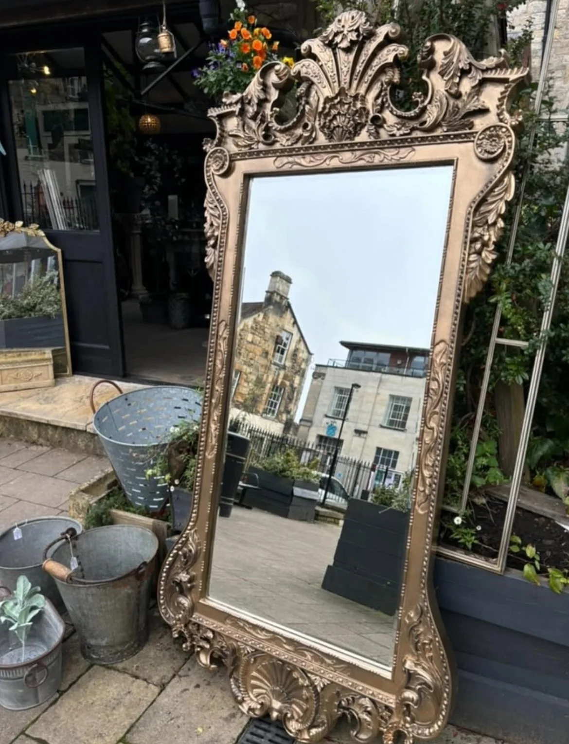An ornate, gold-colored, full-length mirror with decorative carvings on the frame, leaning against a black planter box filled with greenery and flowers; other gardening tools and planters are nearby, and a reflection of buildings and a cloudy sky can be seen in the mirror.