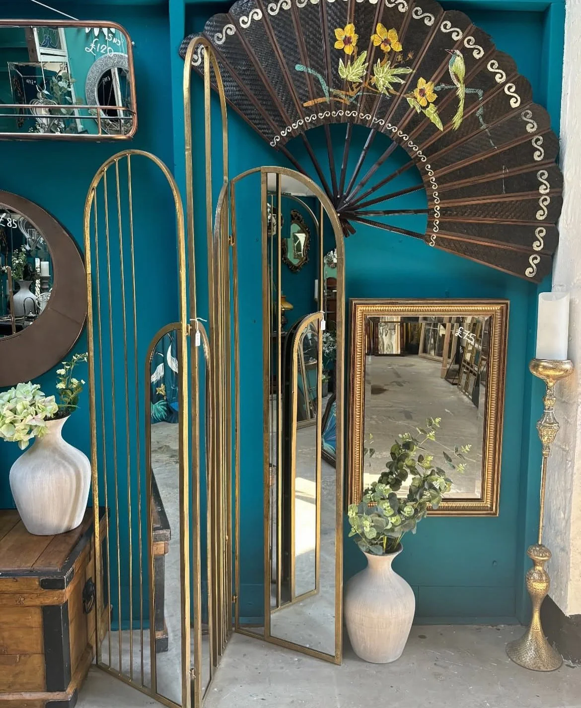 Decorative display with vintage and modern home decor items including vases, mirrors, a folding mirror, a wall fan, and a candlestick, arranged against a teal wall.