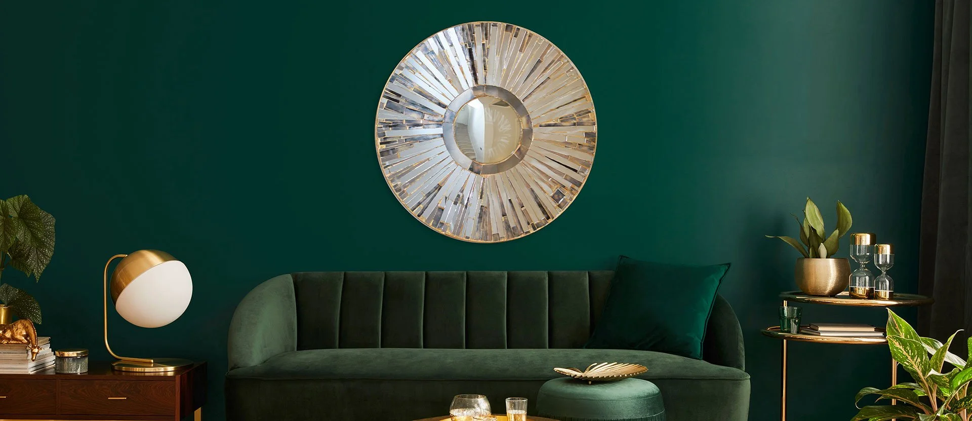 A stylish living room with a dark green wall, a green velvet sofa with a matching pillow, a round side table with gold and glass decor, a modern lamp with a gold base and white shade, and lush green plants. A round decorative mirror with a gold frame hangs on the wall.