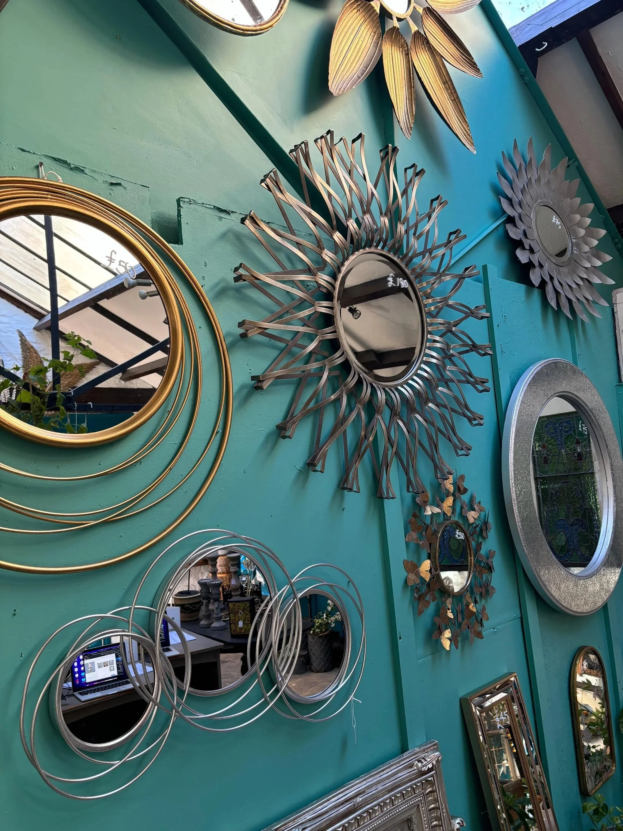 A collection of decorative mirrors with various shapes, sizes, and frame designs on a teal green wall, including circular, floral, and sunburst styles.