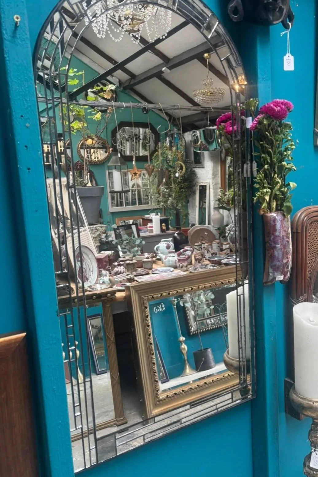 Decorative mirror reflecting an antique store with various vintage items, plants, and chandeliers.