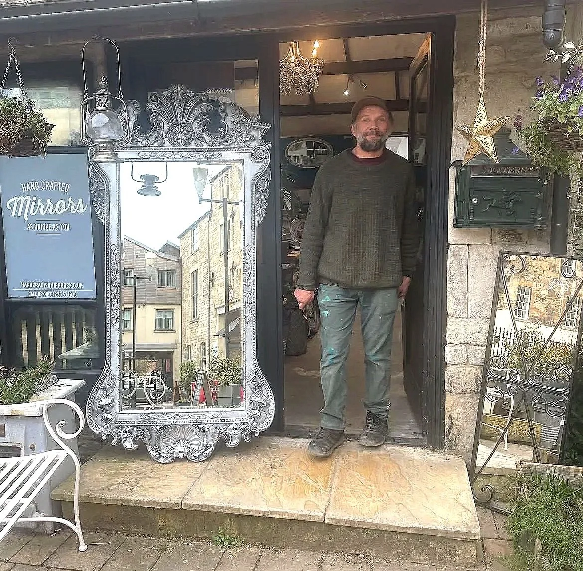A man stands in the doorway of a vintage-style shopfront. The shop has a decorative silver mirror and hanging plants outside, with a sign advertising handcrafted mirrors.
