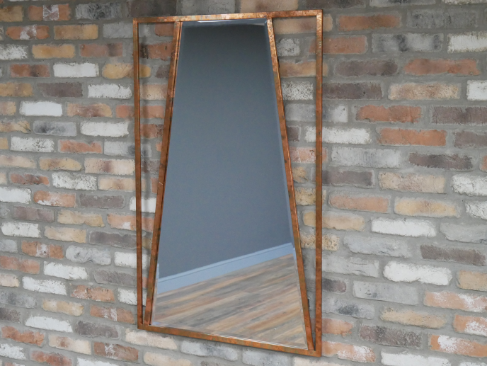 A tall, rectangular mirror with a copper-colored frame is mounted on a brick wall. The mirror reflects a gray wall and a wooden floor.