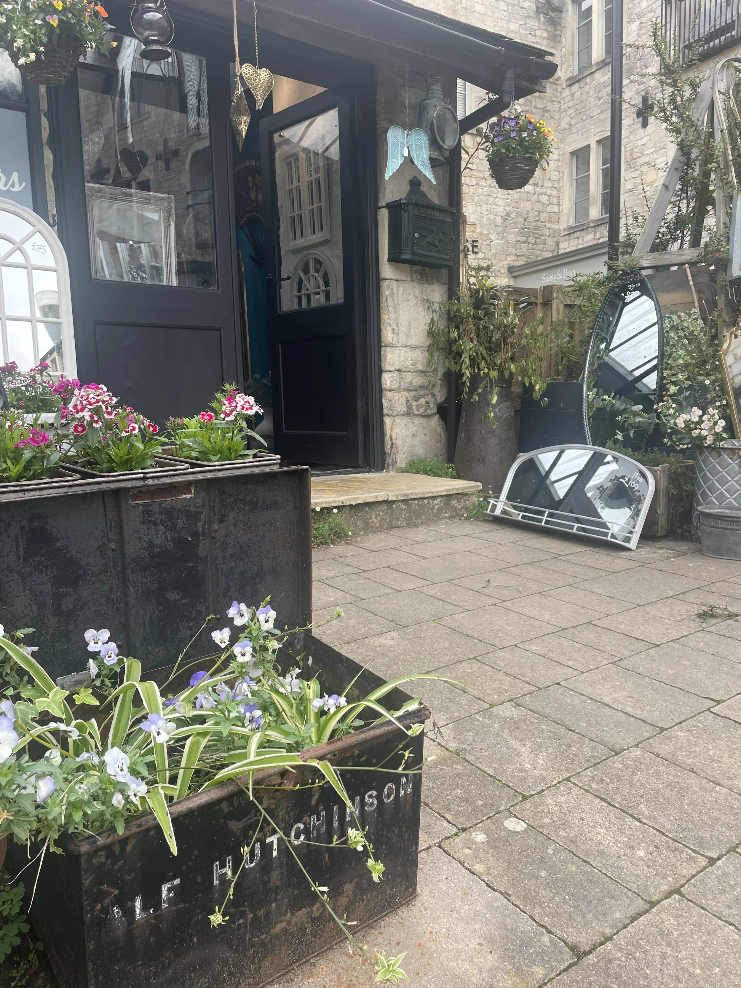 Flower boxes with pink and purple flowers outside a storefront with mirrors, decorative lights, and hanging flower baskets in a stone building setting.