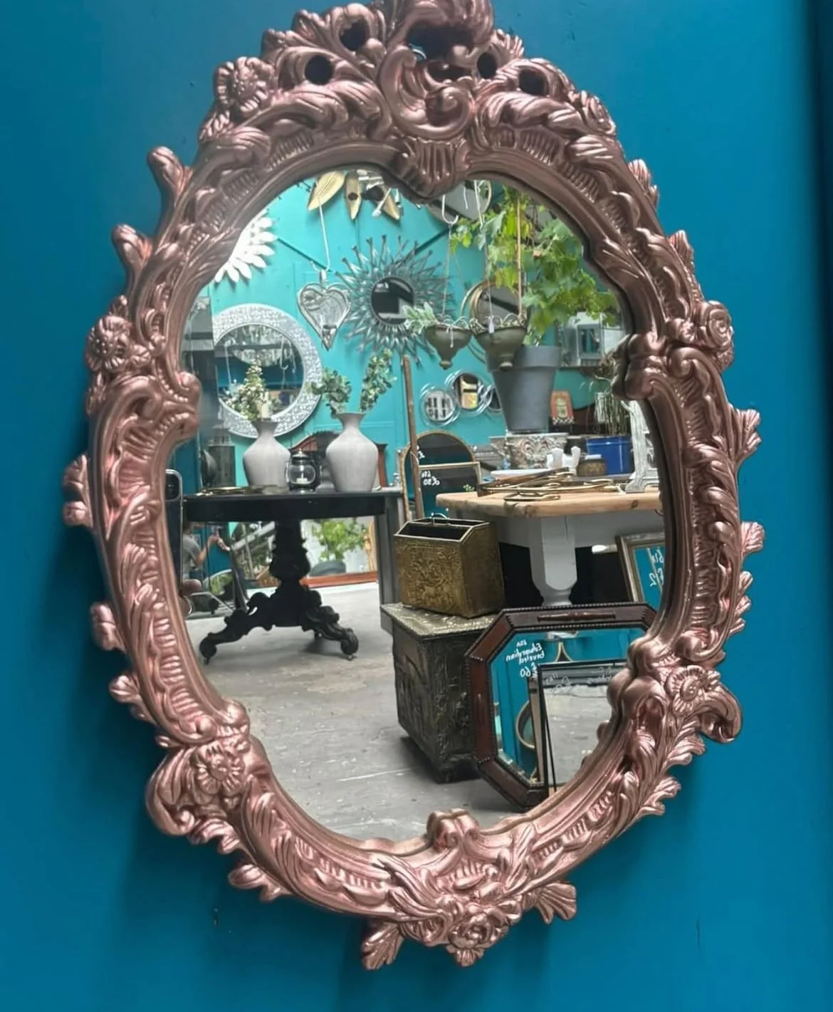 Gold ornate oval mirror with floral details reflecting a room with vases, plants, and decorative items.