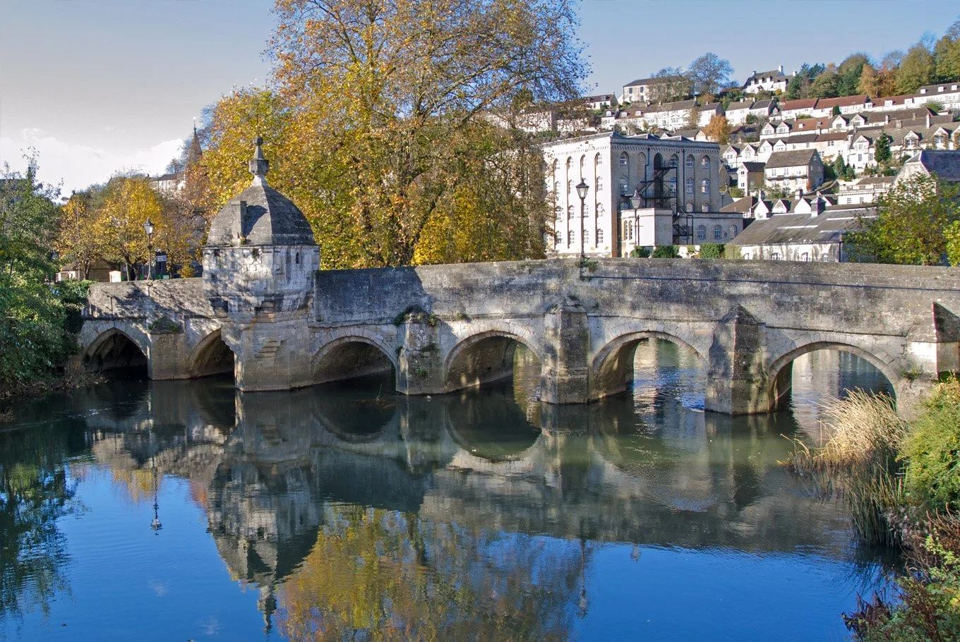 A historic stone bridge over a calm river with autumn trees and residential buildings on a hillside in the background.