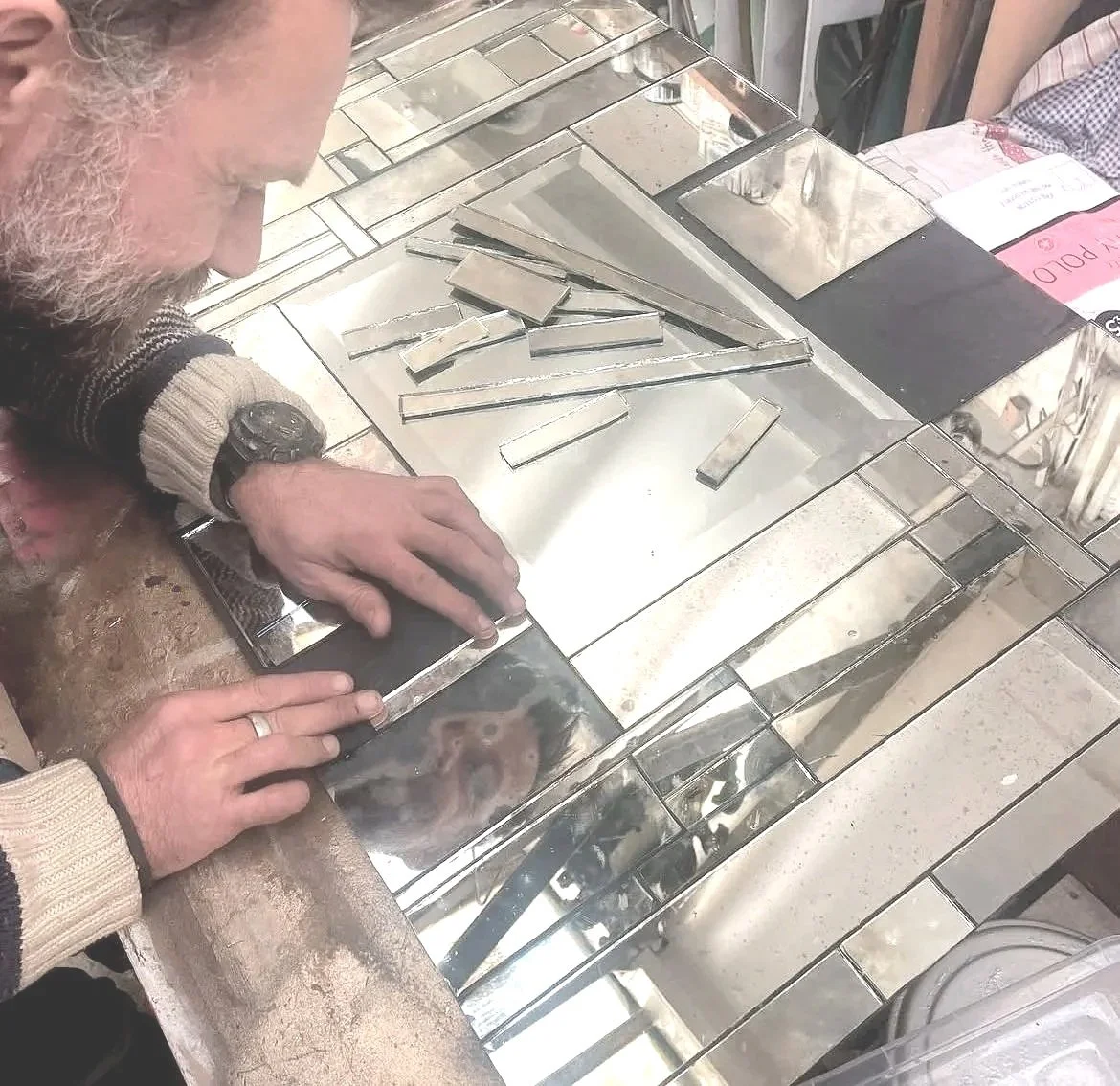 A person with gray hair and a beige sweater is inspecting a collection of small, rectangular mirror tiles arranged on a metallic surface, possibly in a tile store or workshop.