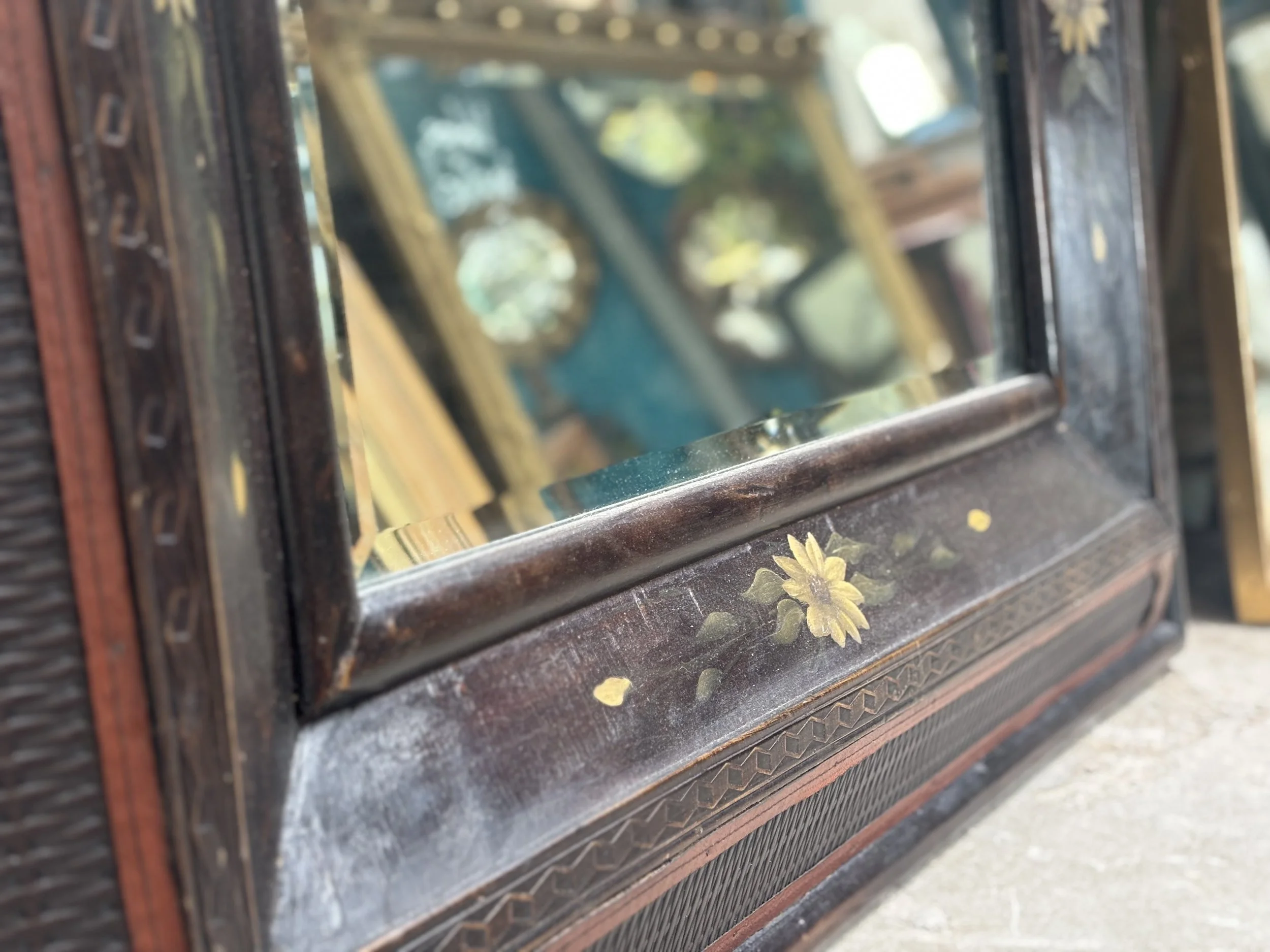 Close-up of an antique wooden framed mirror with a floral painted decoration below the glass, reflecting another mirror with similar floral details.