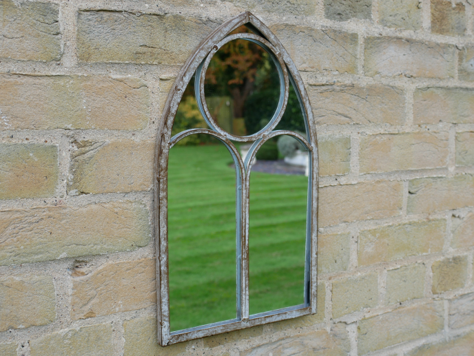 Decorative vintage-style window mirror with a pointed arch design mounted on a brick wall, reflecting a green lawn and outdoor scene.