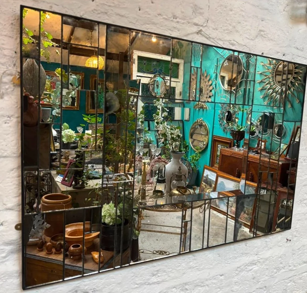 Decorative mirror reflecting an eclectic collection of vintage and antique furniture, artwork, mirrors, plants, and decorative items in a colorful interior space.