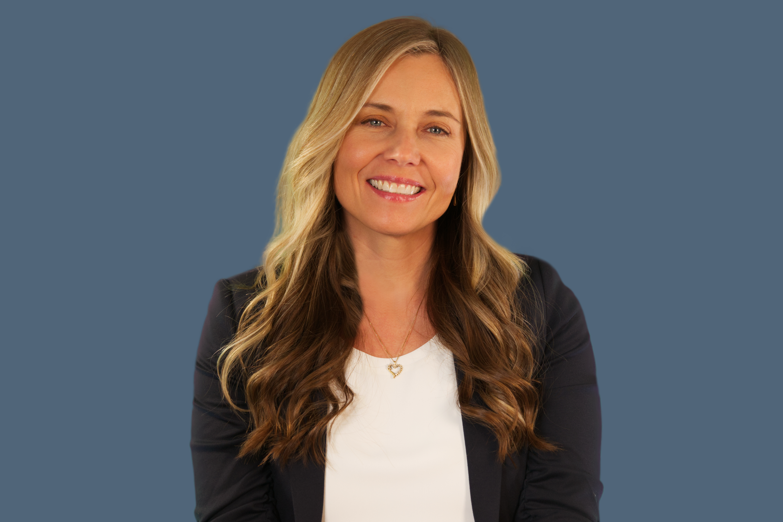 A smiling woman with long, wavy blonde hair, wearing a black blazer over a white top, and a gold necklace with a heart pendant, against a blue background.