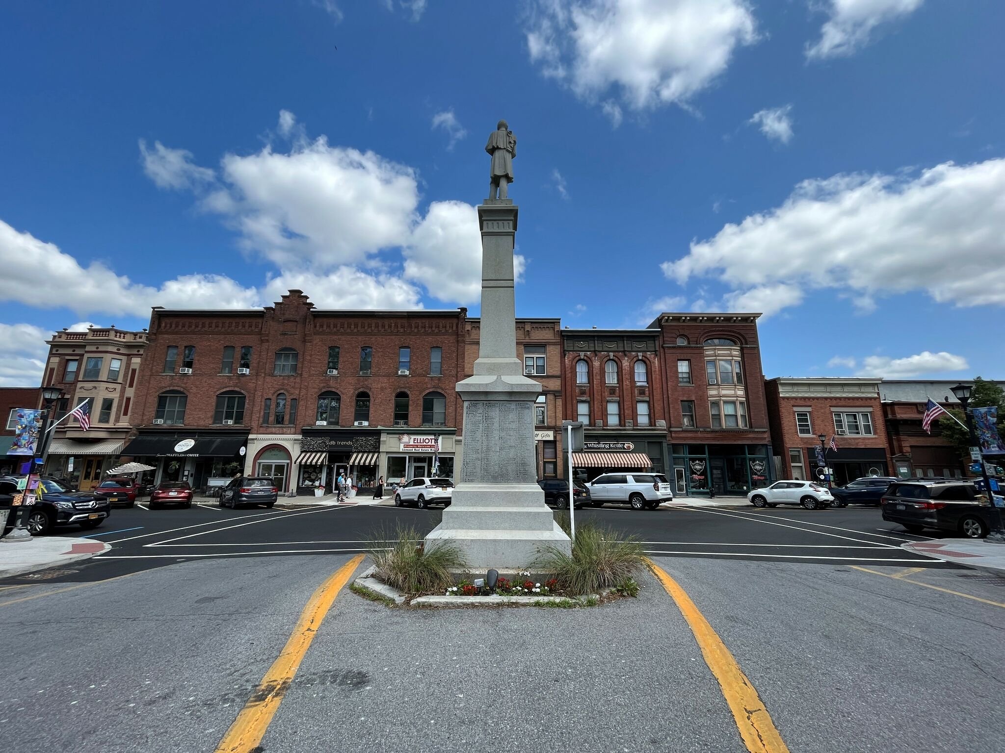A town square with a monument of a soldier on a tall pedestal in the center, surrounded by shops and parked cars beneath a blue sky with white clouds.