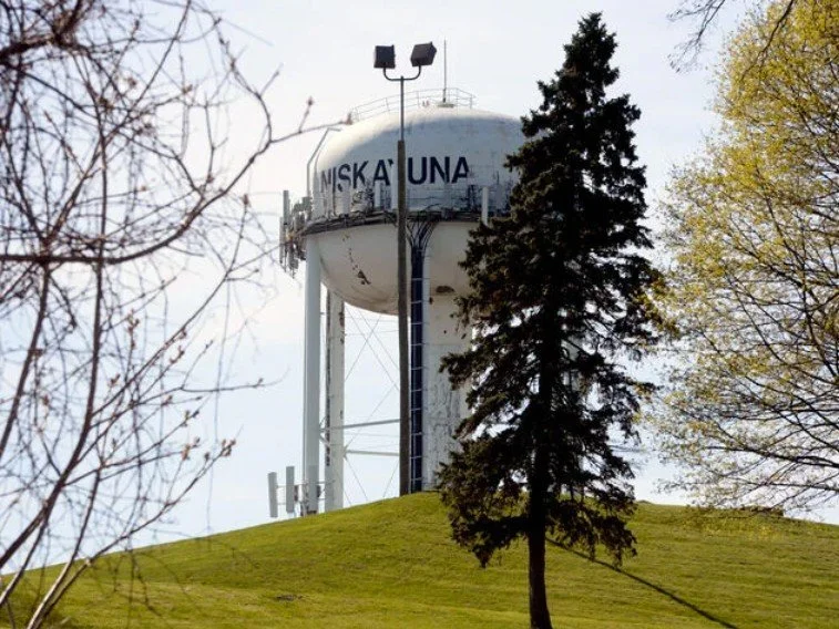 Water tower with 'MISKALUNA' written on it, surrounded by trees and a grassy hill.
