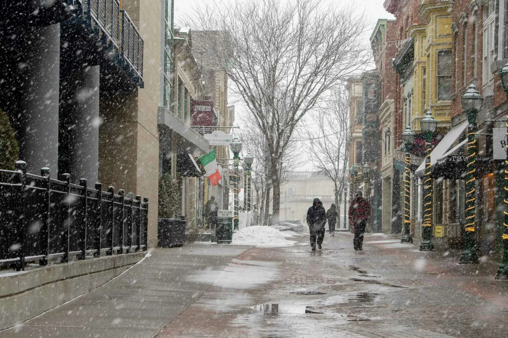 Snow falling on a city street with pedestrians walking, bare trees, and storefronts along both sides, some with holiday lights, during winter.