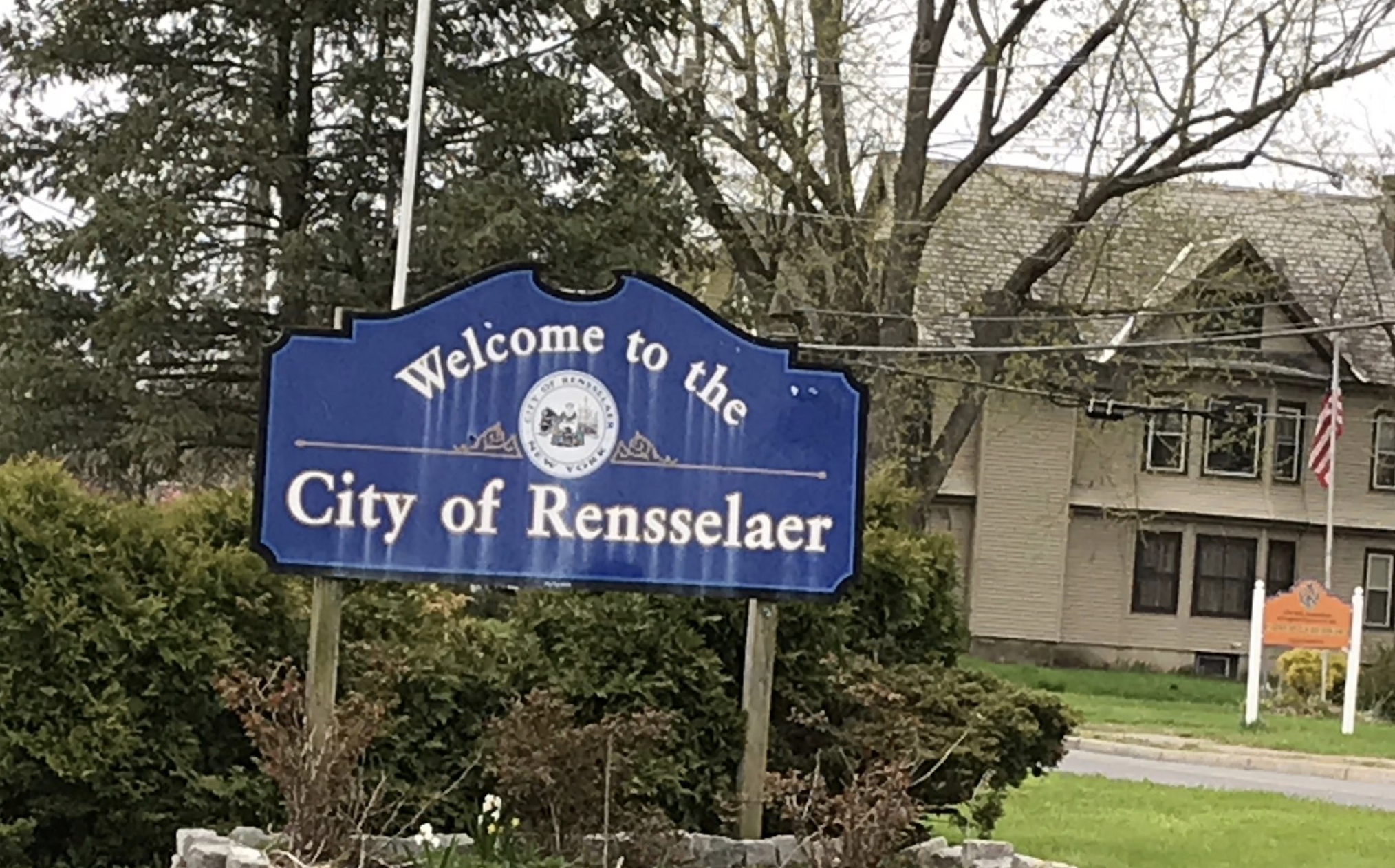 Blue city sign reading 'Welcome to the City of Rensselaer' with trees and residential buildings in the background.
