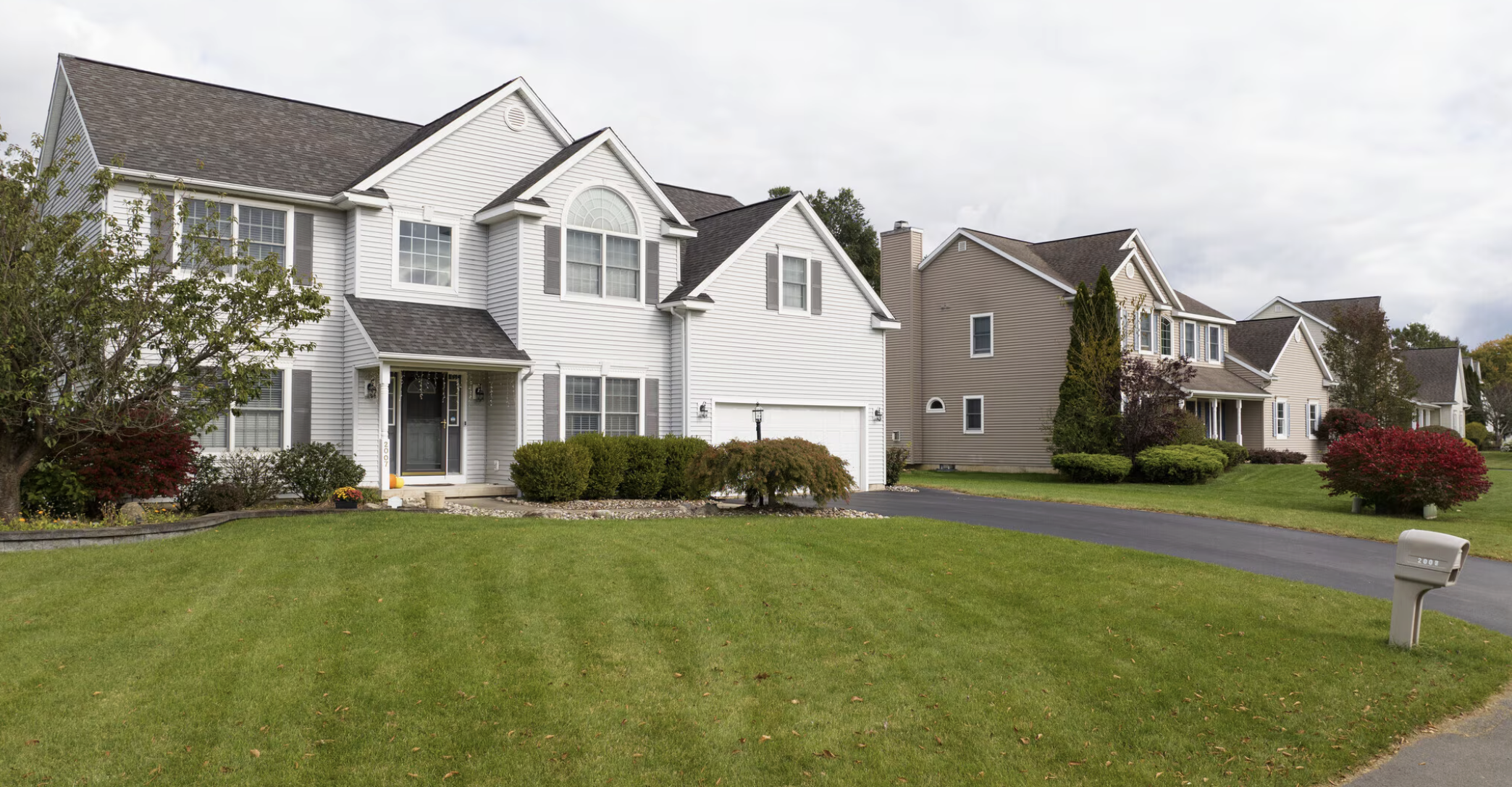 A row of suburban houses with manicured lawns, bushes, trees, and driveways, under a cloudy sky.