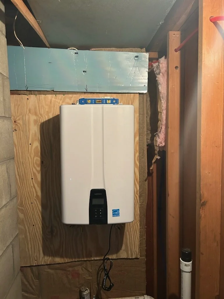 A white tankless water heater installed on a plywood wall in a utility or laundry room. The device is connected to plumbing with exposed pipes and electrical wiring.