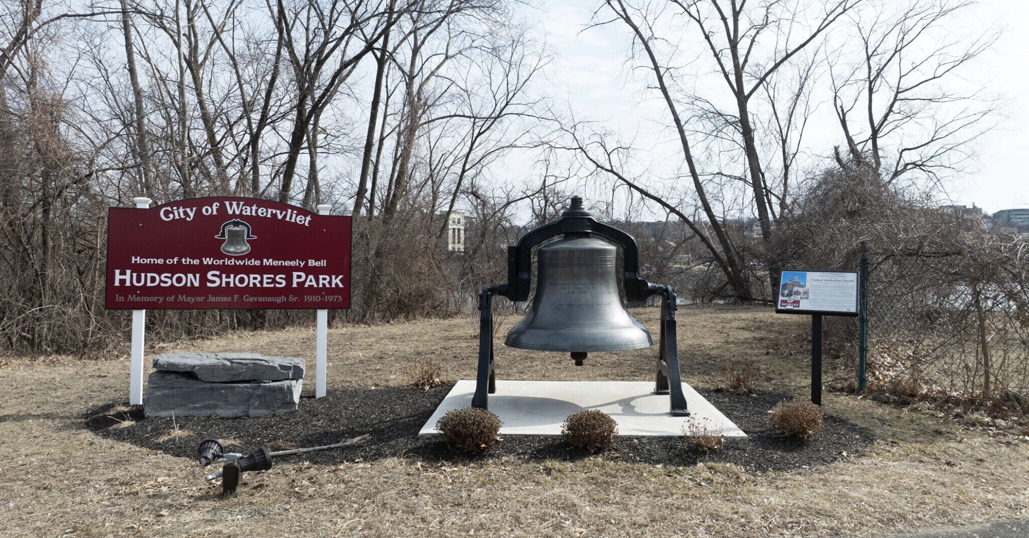 A park entrance sign for Hudson Shores Park in Watervliet, with a large bronze bell on a pedestal in front of a wooded area with bare trees.