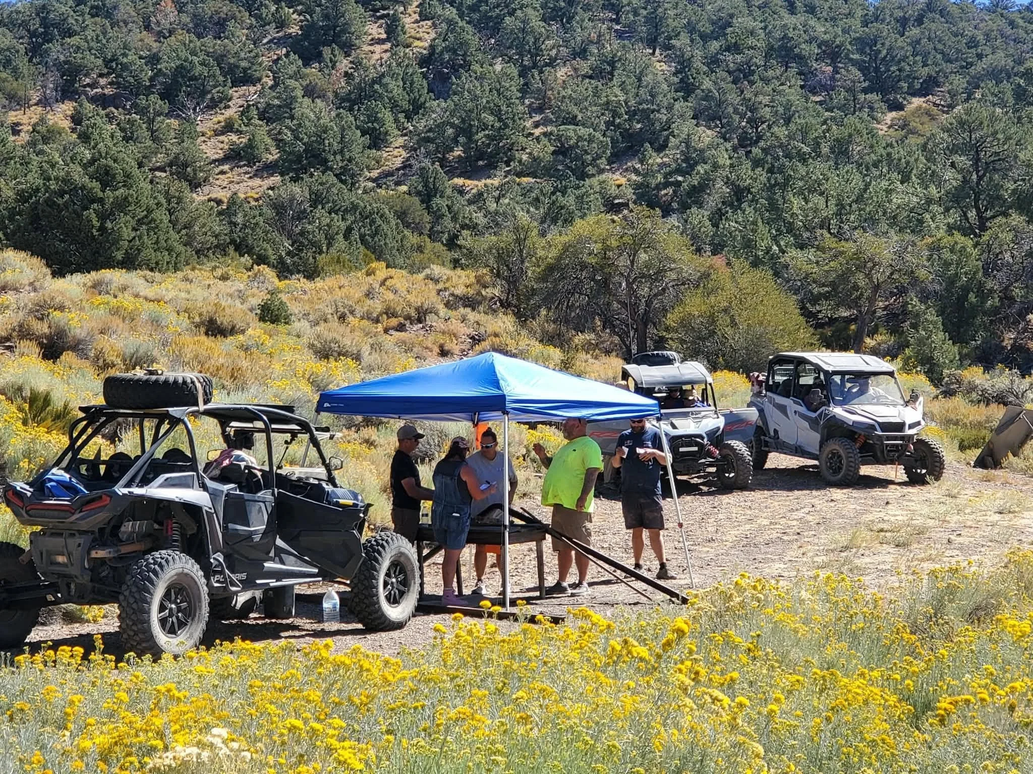 Group of riders gathered under a shade canopy amid blooming desert wildflowers during the 2026 Dyer - Fish Lake Poker Run