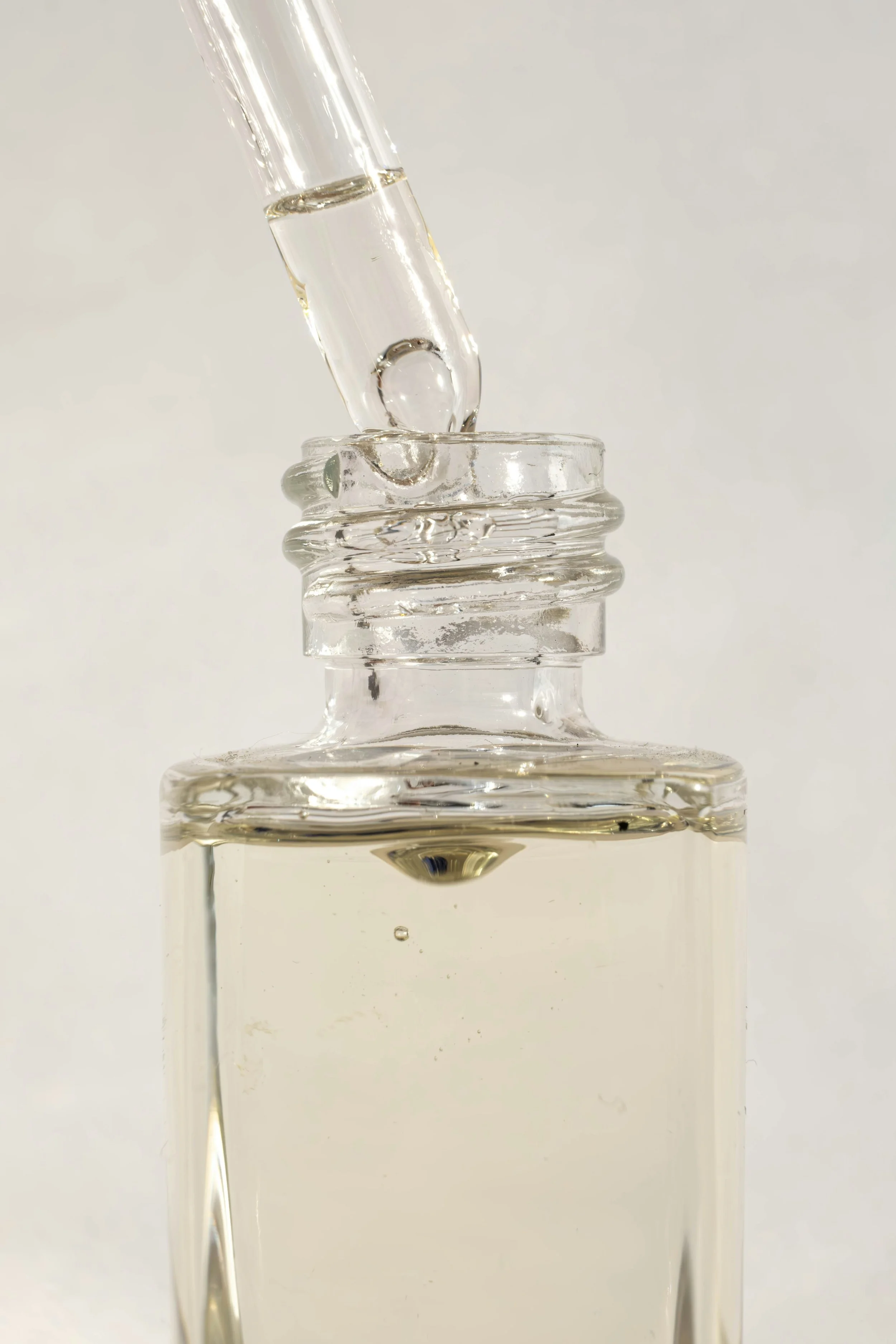 A glass dropper above a glass bottle with a clear liquid, likely a cosmetic or medicinal serum, on a plain white background.