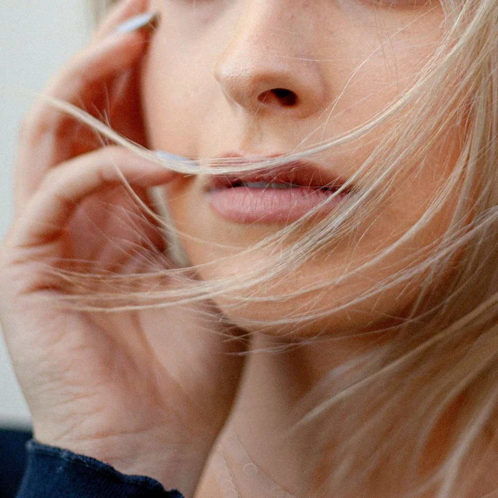 The hand of a white woman, pressed lightly to her face with blonde strands flying across her lips