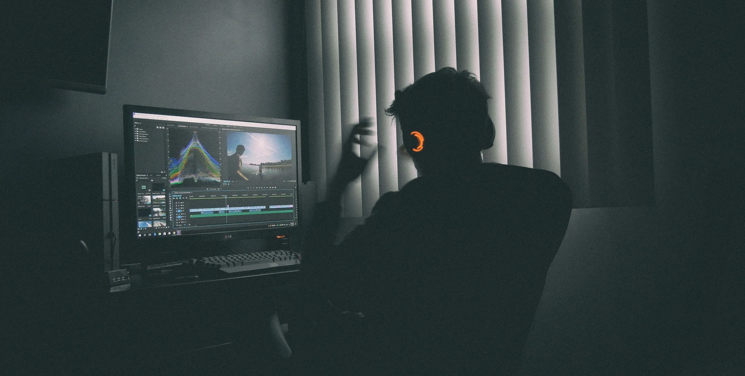 Person wearing headphones working on video editing on a computer in a dark room with vertical blinds.