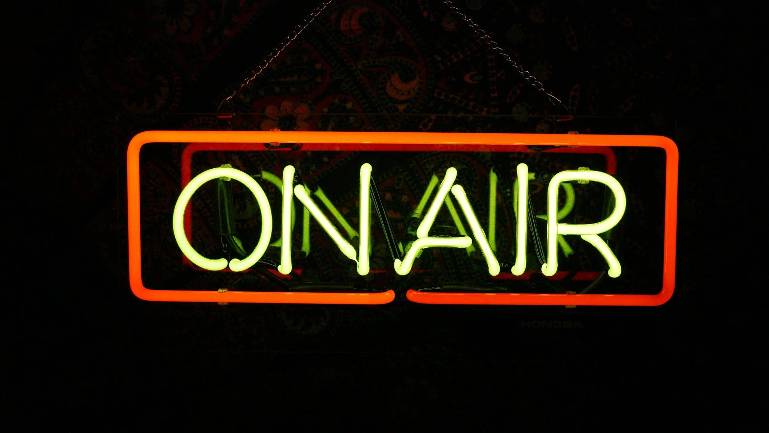 Neon sign reading 'ON AIR' in green and red letters, hanging against a dark background.