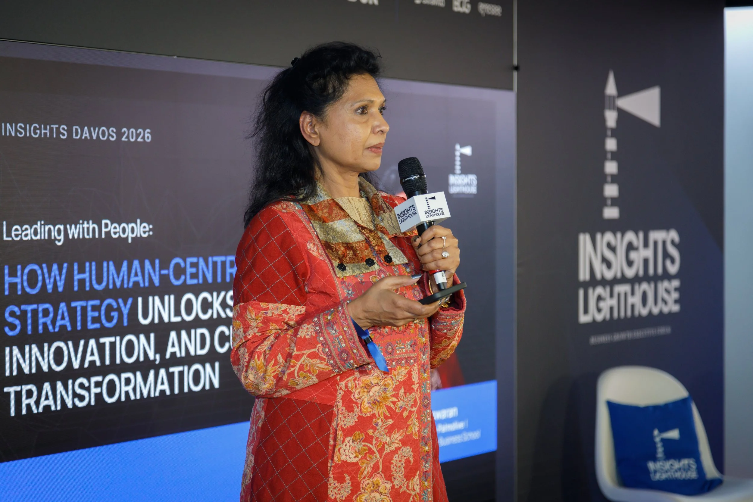 A woman in a red and gold traditional outfit is speaking into a handheld microphone at an event. She is holding a phone in her other hand. Behind her, a screen displays text about Insights Davos 2026 and human-centered strategy. To her right, a banner reads "Insights Lighthouse," and a chair with a blue cushion bearing the event logo is visible.