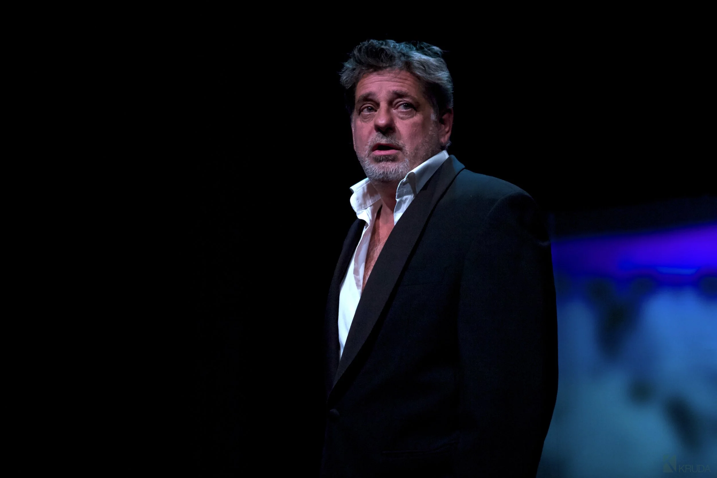 A middle-aged man with gray hair and beard, wearing a black suit and open-collared white shirt, stands on a dark stage with blue lighting in the background, looking serious or concerned.