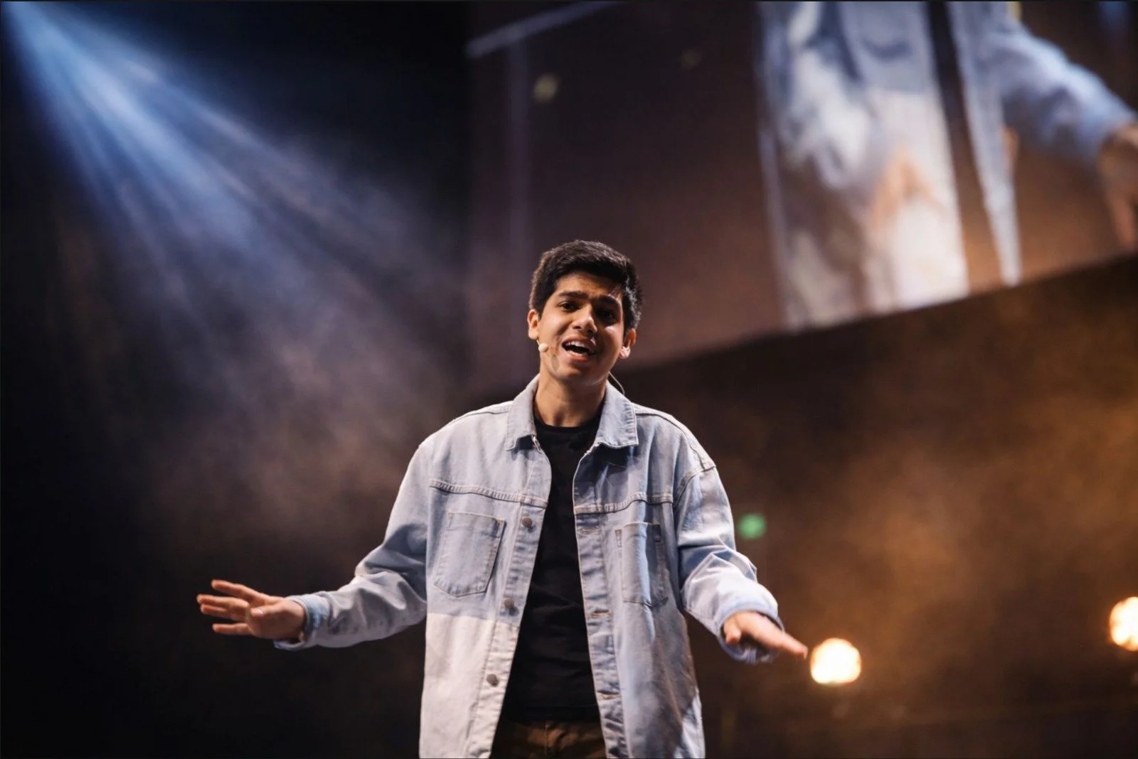 A young man performing on stage, wearing a denim jacket and black shirt, with a microphone headset, gesturing with open hands, and illuminated by stage lights.