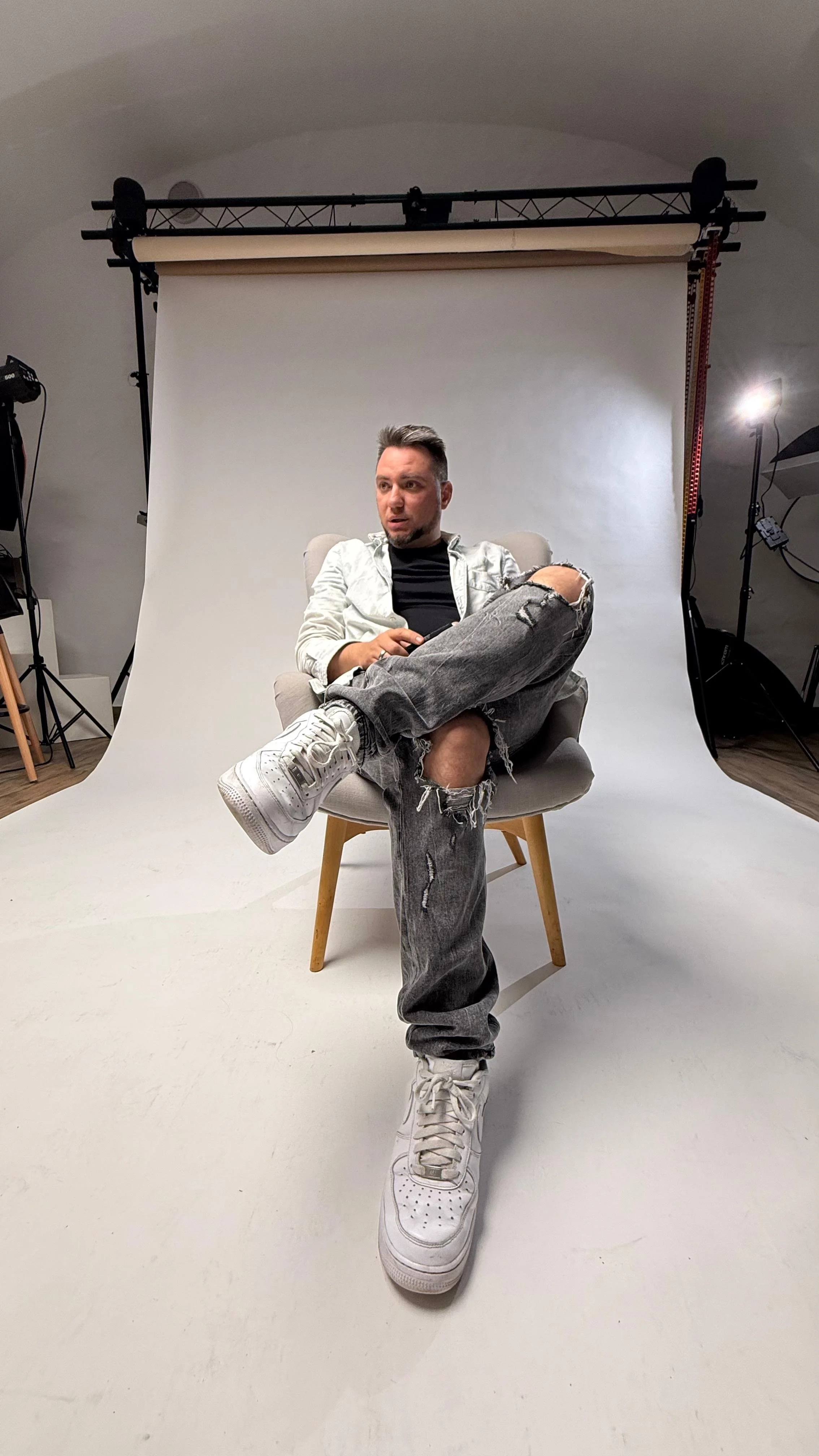 A man sitting on a chair in a photography studio with a white backdrop, wearing ripped jeans, a black shirt, white sneakers, and a light jacket.