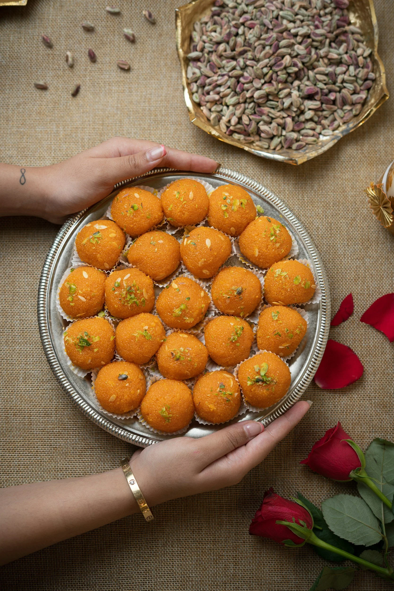 A silver tray holding round, bright orange Indian sweets garnished with chopped almonds and pistachios, placed on a beige woven tablecloth with a bunch of red roses nearby and a bowl of pistachios in the background.