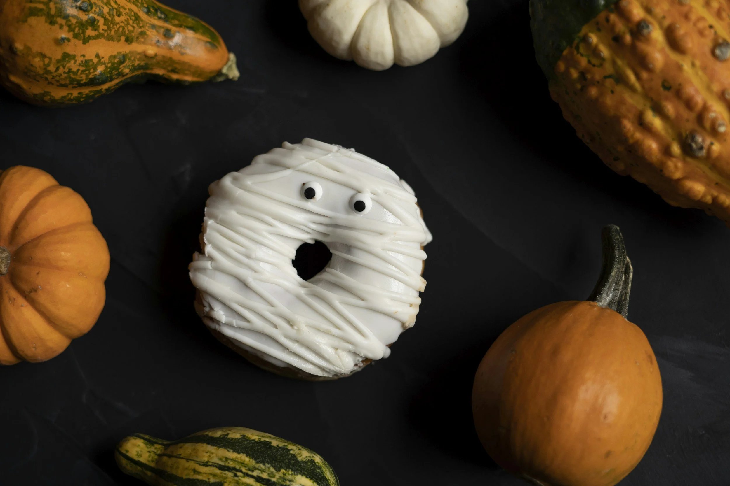 A Halloween-themed donut decorated to look like a mummy with white icing and small candy eyes, surrounded by small pumpkins and gourds on a black background.