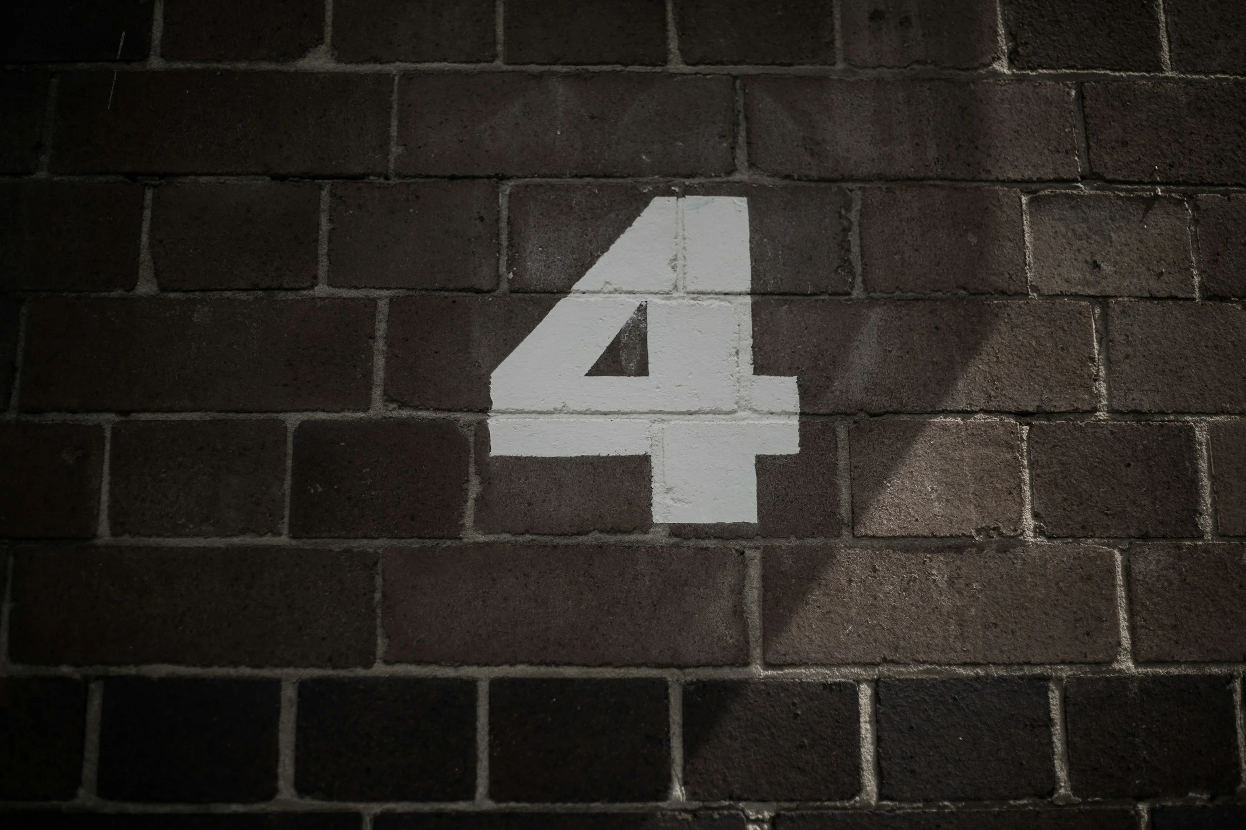 White number 4 painted on a dark brick wall with shadow cast across part of it.