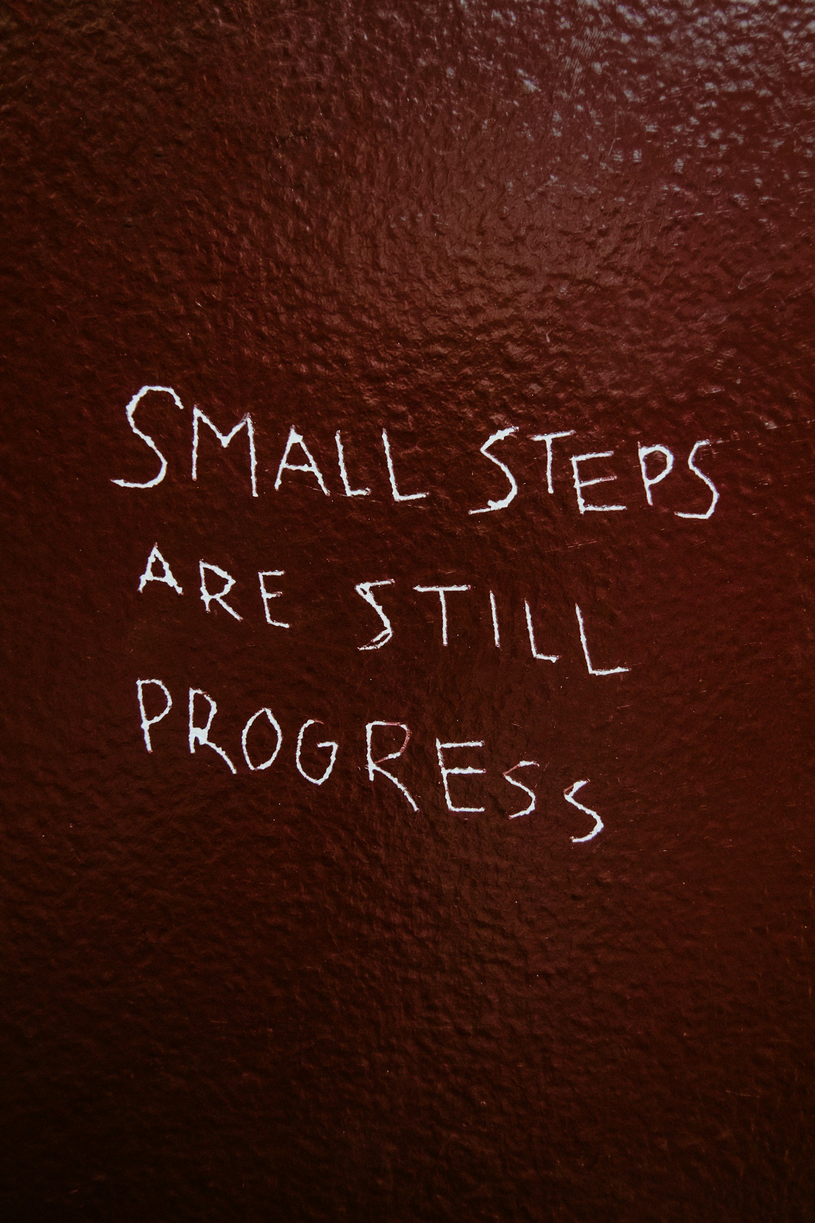 Handwritten sign on a textured red wall that reads, "Small steps are still progress."
