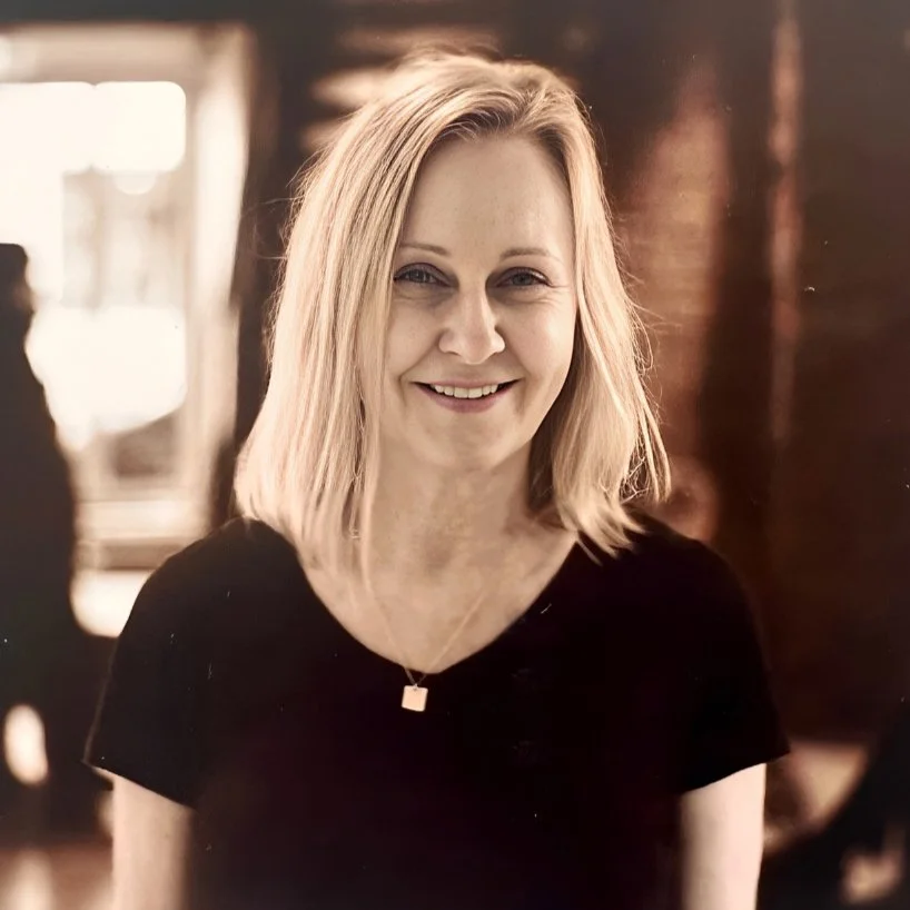 Smiling woman with shoulder-length blonde hair, wearing a black top and a necklace, indoors with warm lighting.
