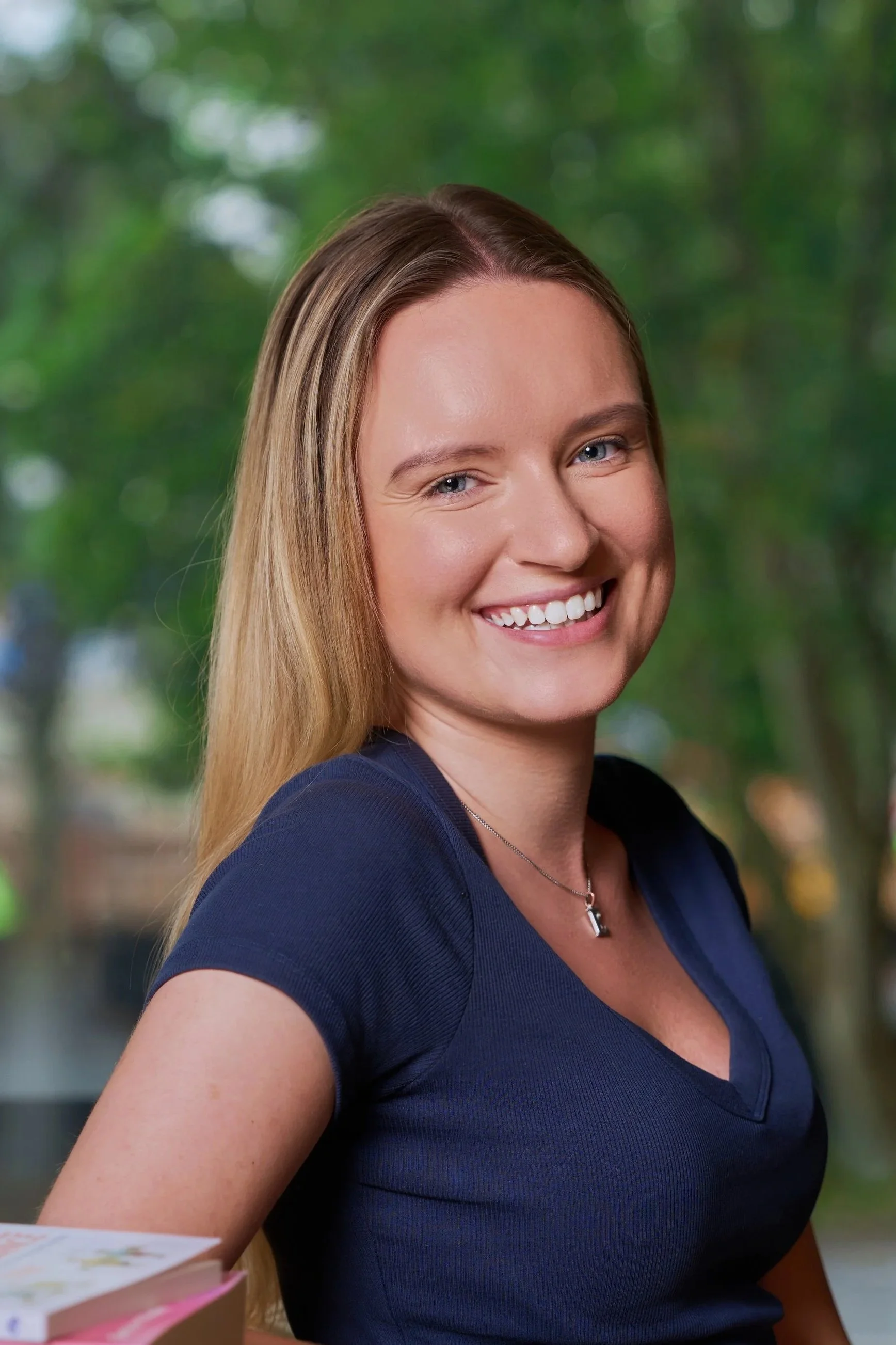 Smiling woman with shoulder-length blonde hair, wearing a black top and a necklace, indoors with warm lighting.