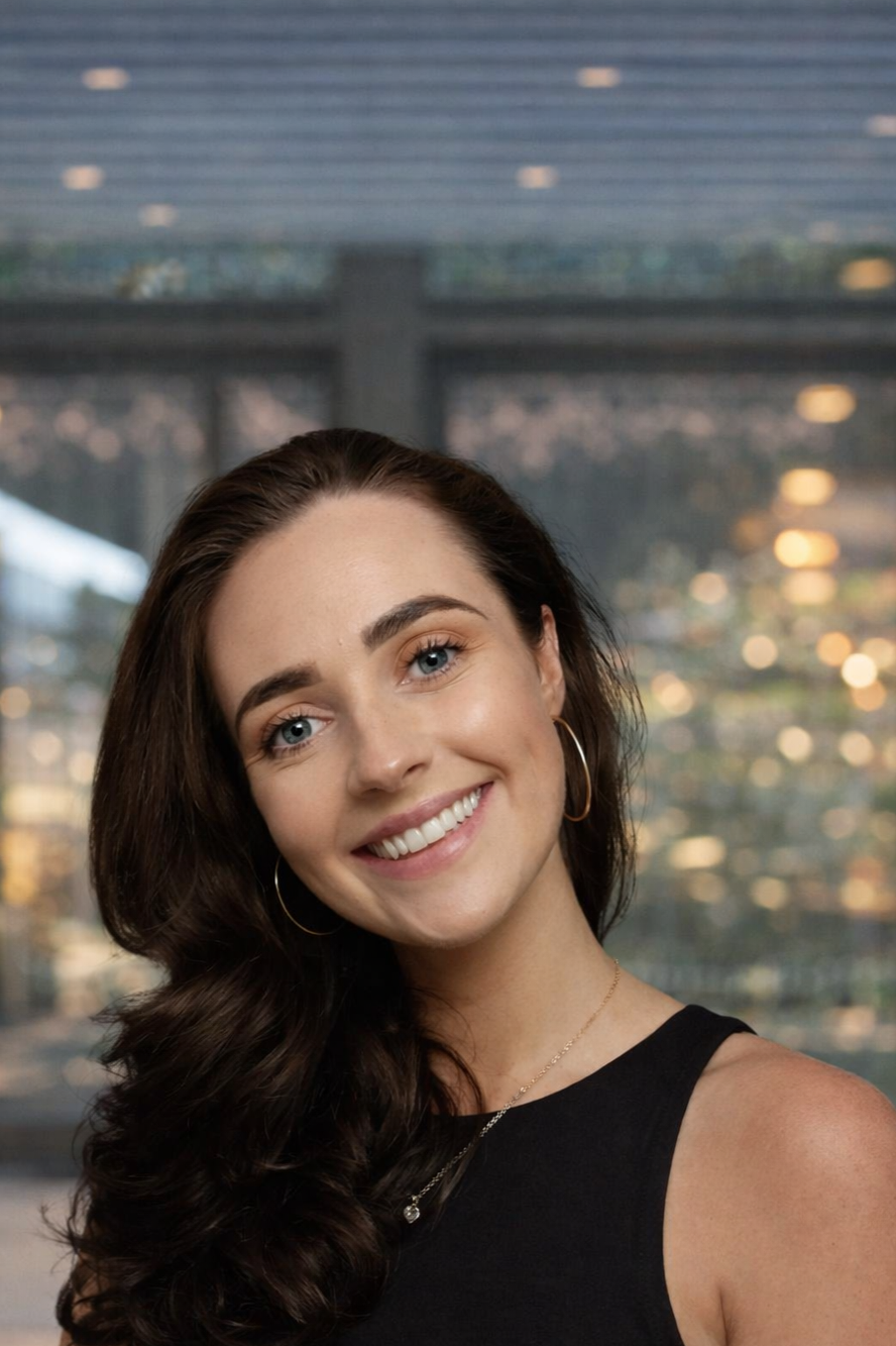 A woman with long dark hair wearing a navy sleeveless top, gold hoop earrings, a gold necklace, and glasses resting on her head, smiling while sitting on a wooden bench with pink cushions in the background.