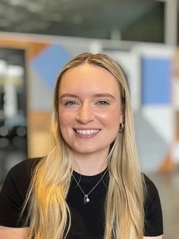 Smiling woman with shoulder-length blonde hair, wearing a black top and a necklace, indoors with warm lighting.